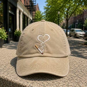 Könnte beinhalten: Eine beigefarbene Baseballkappe mit einer Stickerei auf der Vorderseite. Die Stickerei zeigt eine brennende Zigarette, aus der weißer Rauch aufsteigt und eine Herzform über der Zigarette bildet. Die Kappe hat einen gebogenen Schirm.