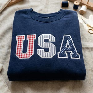Puede incluir: Una sudadera azul marino con la palabra "USA" bordada en la parte delantera. Las letras son de cuadros rojos, rayas azules y blancas y lunares azules. La sudadera está doblada sobre una superficie neutra.