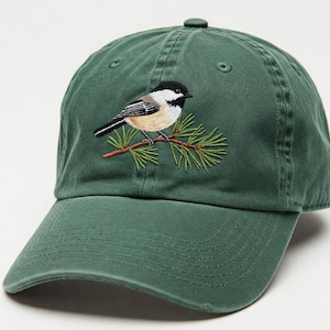 以下が含まれることがあります： 松の枝に止まっている黒頭巾のシジュウカラの刺繍が施された濃い緑色のベースボールキャップです。