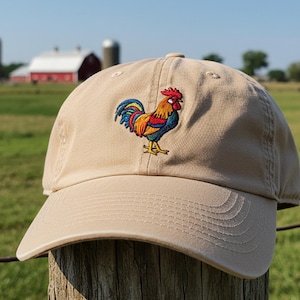 Peut inclure: Une casquette de baseball beige avec un motif de coq brodé. Le coq est coloré avec des accents rouges, orange, jaunes, bleus et verts.