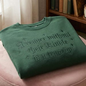 Op de afbeelding: Donkergroene sweatshirt met de geborduurde tekst "A reader without their Kindle is a tragedy". De tekst is in een gotische lettertype. Het sweatshirt ligt op een roze oppervlak.