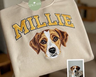 May include: Cream-colored sweatshirt with the name "MILLIE" embroidered in gold lettering above a detailed dog portrait. The dog has brown and white fur and a black nose. A small photo of the dog is in the lower right corner.