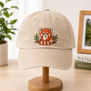Può includere: Un cappellino da baseball beige con un ricamo raffigurante un panda rosso seduto su un ramo con foglie verdi. Il cappellino presenta una corona strutturata, una visiera curva e occhielli cuciti sui pannelli superiori per un look classico.