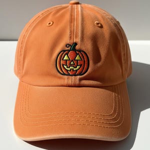 May include: Orange baseball cap with a stitched pumpkin design. The pumpkin has a black outline, a green stem, and a yellow face. The cap has a curved brim and an adjustable strap.