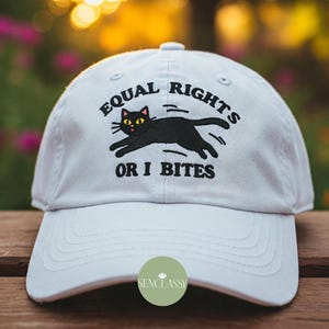 Puede incluir: Gorra de béisbol blanca con bordado negro que dice "Equal Rights or I Bites" y un gato negro con ojos amarillos corriendo por el frente.