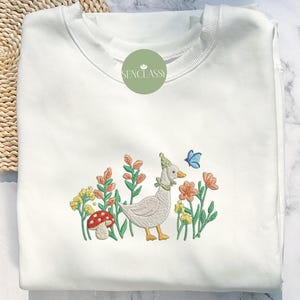 May include: White sweatshirt with an embroidered design of a goose wearing a hat, standing in a field of flowers with a butterfly. The text "Senclassy" is embroidered on the top of the sweatshirt.