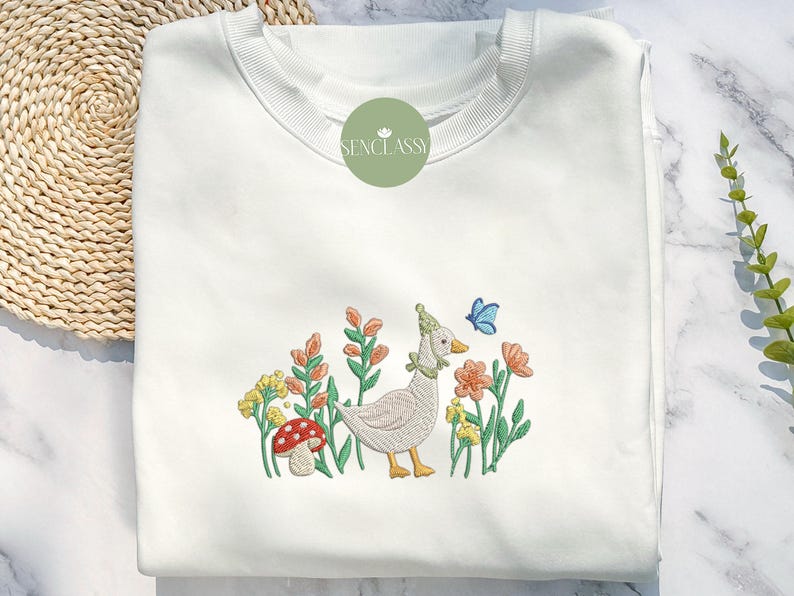May include: White sweatshirt with an embroidered design of a goose wearing a hat, standing in a field of flowers with a butterfly. The text "Senclassy" is embroidered on the top of the sweatshirt.
