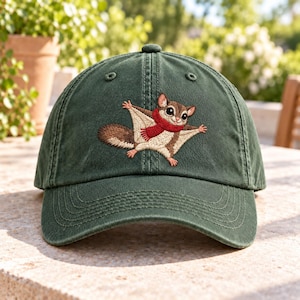 May include: A dark green baseball cap featuring an embroidered design of a brown and cream-coloured flying squirrel wearing a bright red scarf. The cap has a structured crown, a curved brim, and stitched eyelets on the top panels.