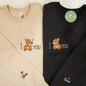 Może przedstawiać: Dwie beżowe i czarne bluzy z nadrukiem kreskówkowego corgi z przodu. Corgi ma serce na plecach i napis "Kocham cię". Bluzy mają nadrukowane imiona "Olivia" i "David" na dole.