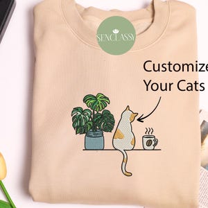 Może przedstawiać: Beżowa bluza z haftowanym wzorem przedstawiającym kota siedzącego obok doniczki z rośliną i filiżanką kawy. Tekst "Customize Your Cats" jest umieszczony nad wzorem.