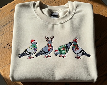 May include: Cream-colored sweatshirt with an embroidered design of four pigeons. Each pigeon is adorned with festive holiday accessories, including Santa hats, reindeer antlers, and scarves. The birds are decorated with Christmas lights and sweaters.