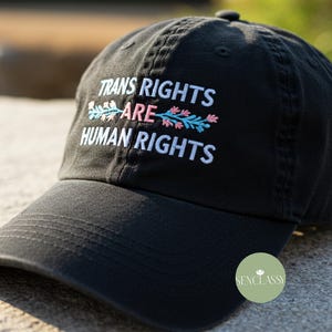 Può includere: Cappellino da baseball nero con ricamo bianco che recita "Trans Rights Are Human Rights" con accenti floreali rosa e blu.