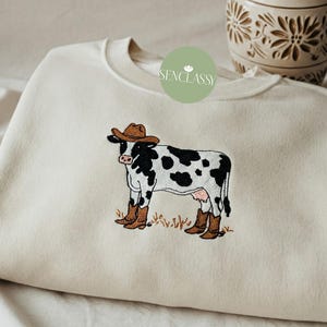 Pode incluir: Uma camisola de cor creme com uma vaca bordada a preto e branco a usar um chapéu de cowboy castanho e botas castanhas. O texto "Senclassy" está bordado a verde na camisola.