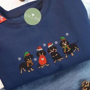 May include: Navy blue sweatshirt featuring an embroidered design of four dachshunds wearing Christmas hats, scarves, and sunglasses. The dogs are surrounded by embroidered snowflakes. The sweatshirt has a crew neck and long sleeves.