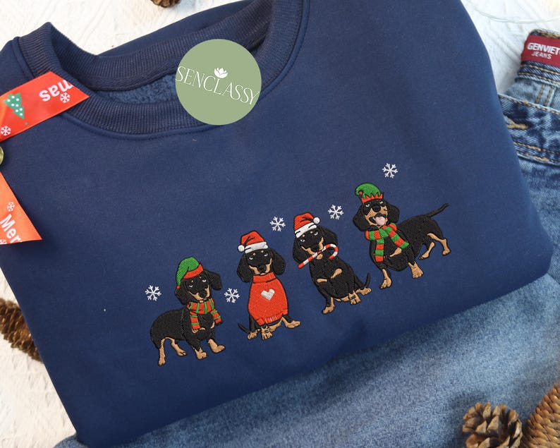 May include: Navy blue sweatshirt featuring an embroidered design of four dachshunds wearing Christmas hats, scarves, and sunglasses. The dogs are surrounded by embroidered snowflakes. The sweatshirt has a crew neck and long sleeves.