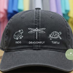 May include: A black baseball cap with a white outline of a frog, dragonfly, and turtle. The words "Frog", "Dragonfly", and "Turtle" are printed below each image.