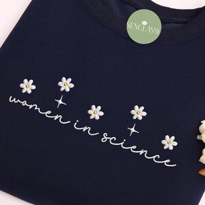 May include: Navy blue sweatshirt with white embroidery that reads "women in science" and features five white daisy flowers with yellow centers.