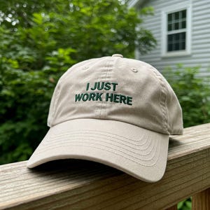Op de afbeelding: Een beige baseballpet met de tekst "I JUST WORK HERE" geborduurd in groen op de voorkant.