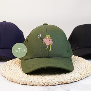 Könnte beinhalten: Eine grüne Baseballkappe mit einem Frosch, der einen rosa Pullover trägt und eine Blume hält. Die Kappe hat einen dunkelblauen Schirm und ein weißes Logo mit der Aufschrift "Senclassy".