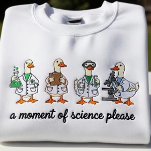 May include: White sweatshirt with an embroidered design of four ducks dressed as scientists, each holding lab equipment. The text "a moment of science please" is embroidered below the ducks.