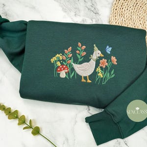 May include: A dark green sweatshirt with an embroidered design of a duck wearing a party hat, surrounded by flowers and a mushroom. The text "Senclassy" is embroidered on the sleeve.