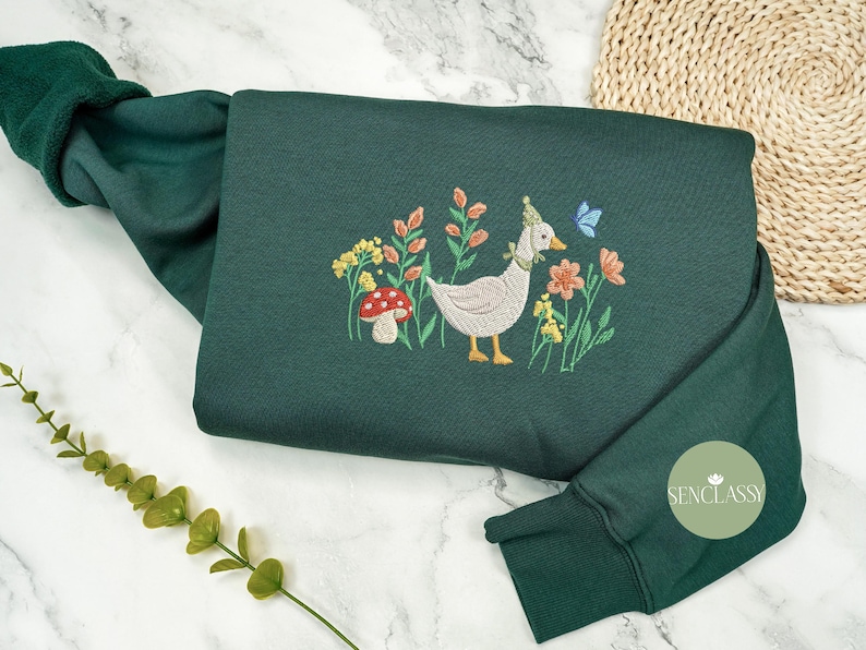 May include: A dark green sweatshirt with an embroidered design of a duck wearing a party hat, surrounded by flowers and a mushroom. The text "Senclassy" is embroidered on the sleeve.