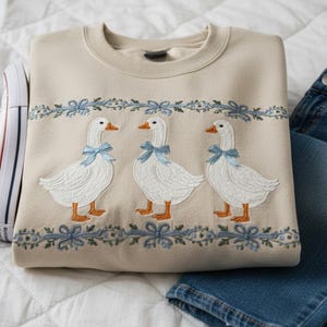 May include: A beige sweatshirt with embroidered white geese, each wearing a blue bow. The geese are set against a blue floral garland. White trainers and blue jeans are also visible in the image. The sweatshirt is folded.