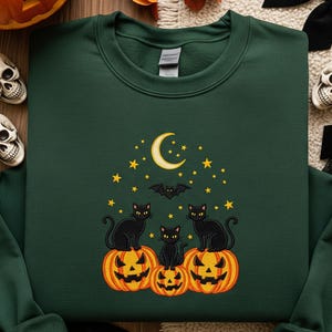 Op de afbeelding: Donkergroene sweatshirt met een geborduurd Halloween-ontwerp. Het ontwerp toont drie zwarte katten die op uitgesneden oranje pompoenen zitten, met een maan, sterren en een vleermuis. De sweatshirt heeft een ronde hals.