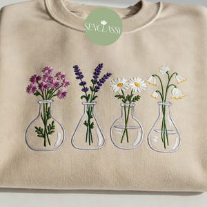 Può includere: Felpa beige con un ricamo floreale. Il design presenta quattro vasi di vetro, ognuno con un fiore diverso: viola, lavanda, margherite e fiori bianchi a forma di campana. La felpa ha un girocollo e maniche lunghe.