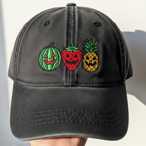 May include: Black baseball cap with embroidered Halloween-themed fruit: a green watermelon, a red strawberry, and a yellow pineapple, each with a jack-o'-lantern face. The cap has a curved brim and a top button.