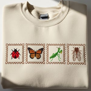 May include: A cream-coloured sweatshirt displaying four insect illustrations within postage stamp frames. The insects depicted are a ladybug, monarch butterfly, praying mantis, and cicada. The illustrations are in red, orange, green, and brown.