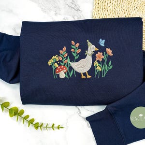 May include: A navy blue sweatshirt with an embroidered design of a goose wearing a party hat, surrounded by flowers and a mushroom. The design is in shades of pink, green, yellow, and white.