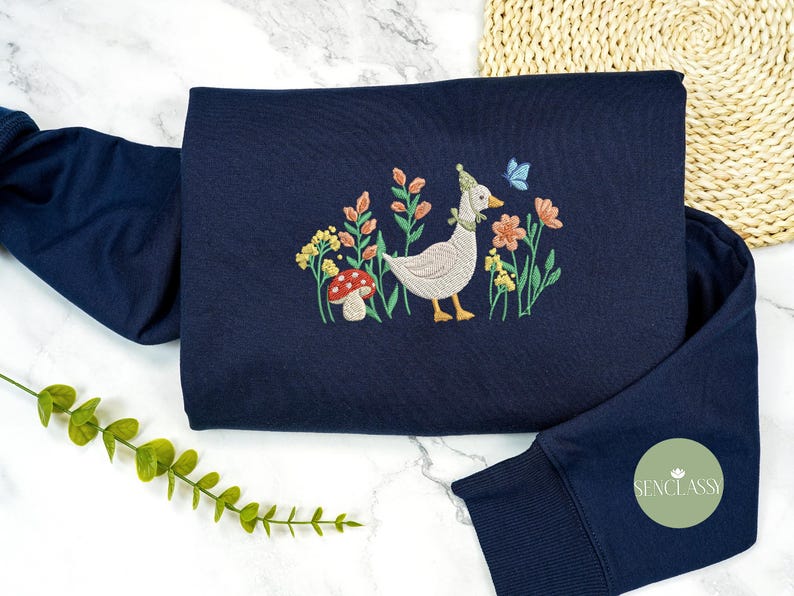 May include: A navy blue sweatshirt with an embroidered design of a goose wearing a party hat, surrounded by flowers and a mushroom. The design is in shades of pink, green, yellow, and white.