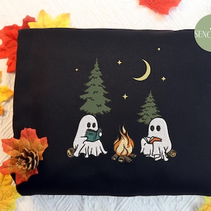May include: Black sweatshirt with an embroidered design of two ghosts reading books by a campfire in a forest. The design includes a crescent moon and stars.