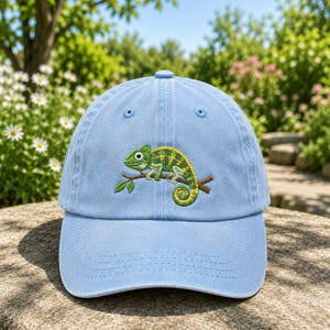 Può includere: Un cappellino da baseball azzurro con un ricamo raffigurante un camaleonte verde e giallo appollaiato su un ramo di un albero marrone con piccole foglie verdi. Il cappellino presenta una corona strutturata e una visiera curva.