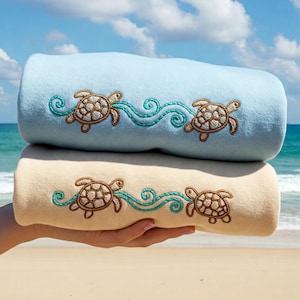 Puede incluir: Dos toallas dobladas con tortugas marinas y olas bordadas. La toalla de arriba es azul claro y la toalla de abajo es crema.