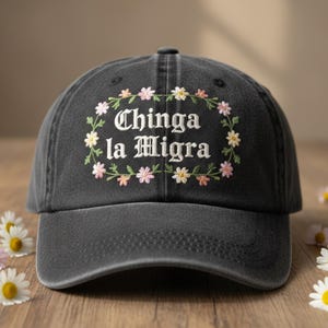 Può includere: Un cappellino da baseball nero con la scritta ricamata "Chinga la Migra" circondata da una ghirlanda floreale bianca, rosa e verde. Il cappellino è appoggiato su una superficie di legno con margherite.