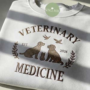 Pode incluir: Camisola branca com bordado castanho que diz "Veterinary Medicine" com um desenho de cão, gato e pássaro. O texto "Est. 2024" também está bordado na camisola.