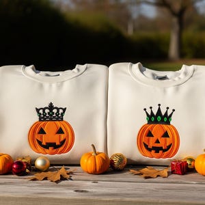 Puede incluir: Dos sudaderas color crema con diseños de calabazas bordadas, cada una con una corona negra. Las calabazas son naranjas con caras negras. Pequeñas calabazas, hojas de otoño y adornos están dispuestos frente a las sudaderas sobre una superficie de madera.