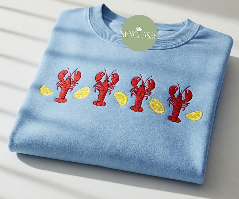 May include: A light blue sweatshirt with embroidered red lobsters and yellow lemon slices. The design features four lobsters and lemon slices. The sweatshirt has a crew neck and is folded neatly. The brand name "SENCLASSY" is on the top right.