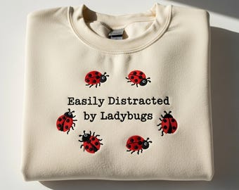 May include: Cream-colored sweatshirt with the embroidered text "Easily Distracted by Ladybugs." Six red ladybugs with black spots and black heads are embroidered around the text. The sweatshirt is folded.