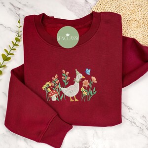 May include: A maroon sweatshirt with an embroidered design of a goose wearing a blue hat and a butterfly, surrounded by flowers and a mushroom.