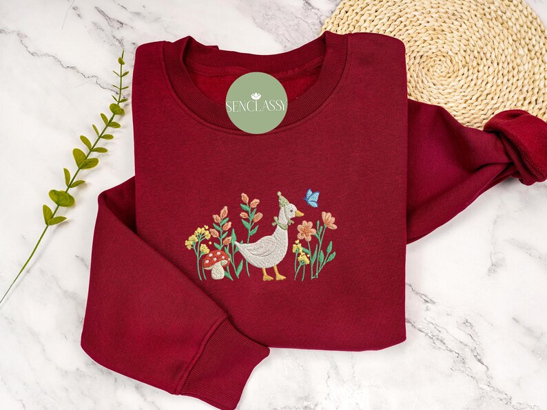 May include: A maroon sweatshirt with an embroidered design of a goose wearing a blue hat and a butterfly, surrounded by flowers and a mushroom.