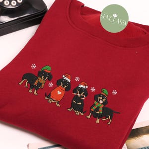 May include: A red sweatshirt featuring an embroidered design of four dachshunds in festive holiday attire. Each dog wears a hat and scarf, with snowflakes scattered around them. The sweatshirt has a crew neck.