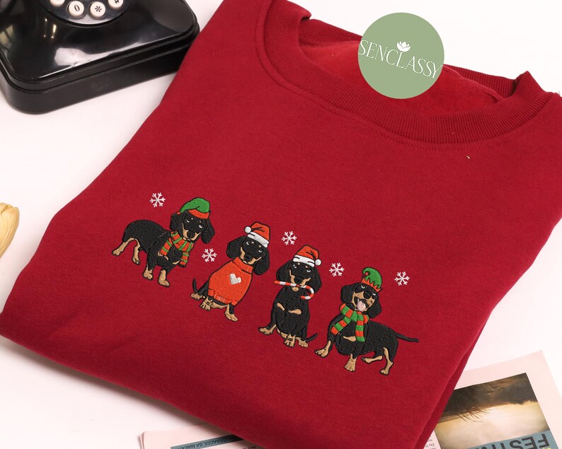 May include: A red sweatshirt featuring an embroidered design of four dachshunds in festive holiday attire. Each dog wears a hat and scarf, with snowflakes scattered around them. The sweatshirt has a crew neck.