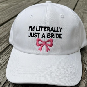 May include: White baseball cap with the embroidered text "I'M LITERALLY JUST A BRIDE" in black, and a pink bow. The cap has a curved brim and a button on top. The background is a weathered wooden surface.