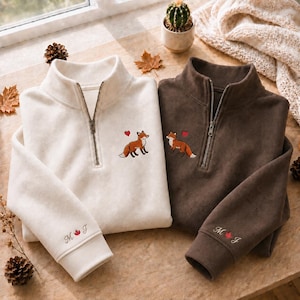 Könnte beinhalten: Ein cremefarbener Pullover mit Viertelreißverschluss und ein dunkelbrauner Pullover mit Viertelreißverschluss, beide mit einem gestickten Fuchs und rotem Herz auf der Brust sowie den Buchstaben M und J mit rotem Ahornblatt am Ärmelbündchen.