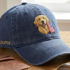 May include: A blue baseball cap with an embroidered golden retriever holding a small American flag in its mouth. The word "COOPER" is embroidered in gold on the side of the cap. The cap is made of a soft, worn material.