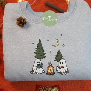 May include: A light blue sweatshirt with a white embroidered design of two ghosts reading books by a campfire in the woods. The text "Senclassy" is embroidered on the top left corner of the sweatshirt.