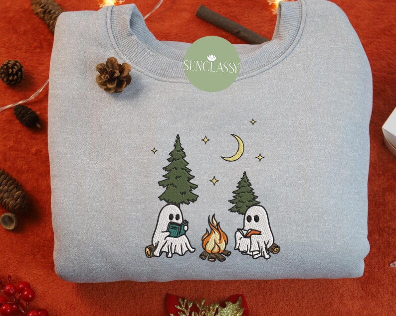 May include: A light blue sweatshirt with a white embroidered design of two ghosts reading books by a campfire in the woods. The text "Senclassy" is embroidered on the top left corner of the sweatshirt.